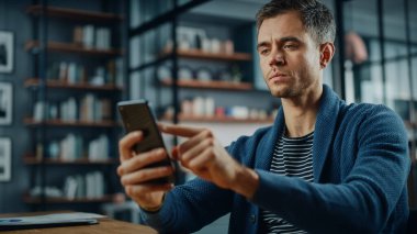 Stil Oturma Odası 'ndaki masada otururken akıllı telefon kullanan ciddi bir beyaz adam. Evde Gülümseyen ve İnternet üzerinden İş Arkadaşları ve Müşterilerle Sohbet eden Genç Adam. Sosyal ağlar kullanılıyor.