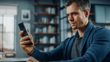 Stil Oturma Odası 'ndaki masada otururken akıllı telefon kullanan ciddi bir beyaz adam. Evde Gülümseyen ve İnternet üzerinden İş Arkadaşları ve Müşterilerle Sohbet eden Genç Adam. Sosyal ağlar kullanılıyor.