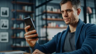 Stil Oturma Odası 'ndaki masada otururken akıllı telefon kullanan ciddi bir beyaz adam. Evde Gülümseyen ve İnternet üzerinden İş Arkadaşları ve Müşterilerle Sohbet eden Genç Adam. Sosyal ağlar kullanılıyor.