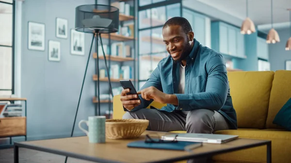 英俊的非洲裔美国人坐在舒适客厅的沙发上 一边用智能手机 自由职业者在家里工作 浏览互联网 使用社交网络 享受平淡的乐趣 — 图库照片