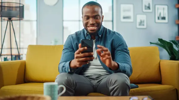 兴奋的非洲裔美国人一边在客厅里坐在沙发上一边用智能手机 快乐的男人在家里微笑 在网上与同事和客户聊天 使用社交网络 — 图库照片