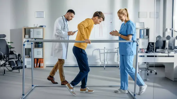 Hastane Fizik Terapisi: Yaralı Güçlü Hasta İlk Adımlarını Yapıyor, Paralel Barlar İçin Yürüyor. Fizyoterapist, Rehabilitasyon Doktoru Yardım Engelli Kişi.