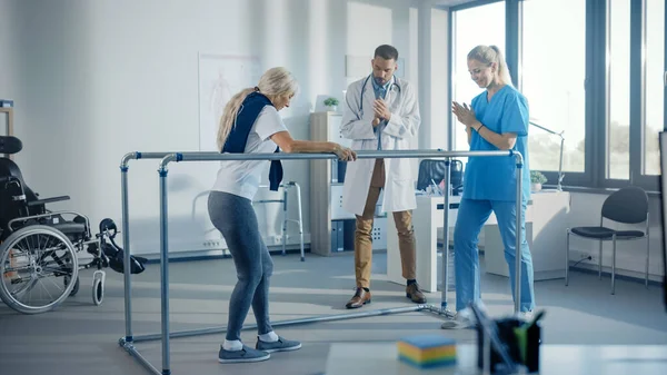 Hospital Physical Therapy: Strong Senior Female Disabled Patient with Injury Making First Steps, Walks Holding for Parallel Bars. Physiotherapist, Rehabilitation Doctor Applaud, Encourage Old Woman