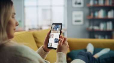 Güzel Otantik Kadın Teknoloji Hakkında Son Haberleri ve Makaleleri Okumak İçin Akıllı Telefon Kullanıyor. Ev Oturma Odası 'nda bir koltukta yatıyor. Evden Çalışan Serbest Çalışan, İnternette Sörf Yapan.