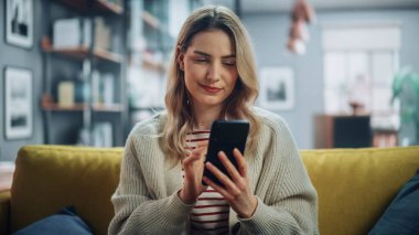 Şık Oturma Odası 'nda rahat bir koltukta dinlenirken akıllı telefon kullanan güzel beyaz kadın. Evde Genç Bir Kadın, İnternet Taraması, Sosyal Ağ Kullanımı, Daire 'de Eğlenme.