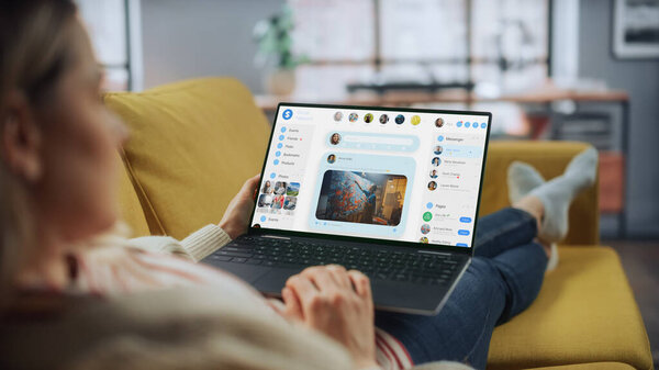 Красивая кавказская женщина использует ноутбук для общения с дьяволами в социальной сети в гостиной, лежа на диване. Маркетолог социальных сетей, работающий из дома, серфинг в Интернете.