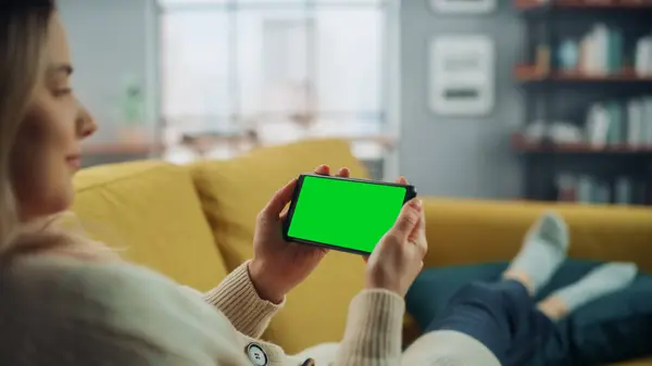 漂亮的正宗女人躺在沙发沙发上 用装有绿色屏幕的智能手机在家里客厅里展示 她正在浏览互联网 并在社交网络上查看视频 — 图库照片