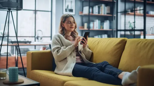 漂亮的白人女性在时髦的客厅里使用智能手机 在舒适的沙发沙发上休息 在家里的年轻女人 浏览互联网 利用社交网络 平易近人 — 图库照片