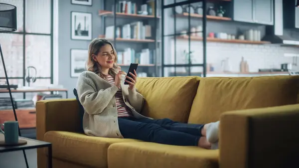 漂亮的白人女性在时髦的客厅里使用智能手机 在舒适的沙发沙发上休息 在家里的年轻女人 浏览互联网 利用社交网络 平易近人 — 图库照片