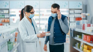 Covid-19 Yüz Maskesi Giyen Eczane: Profesyonel Eczacı Yakışıklı Tıbbi Tavsiyeli Erkek Müşteriye Yardım Ediyor, Konuşuyor. İlaç, hap, sağlık hizmeti, spor ürünleriyle dolu bir eczane.