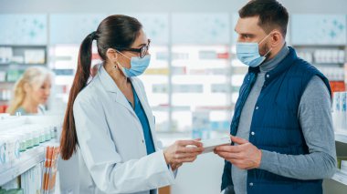 Covid-19 Yüz Maskesi Giyen Eczane: Profesyonel Eczacı Yakışıklı Tıbbi Tavsiyeli Erkek Müşteriye Yardım Ediyor, Konuşuyor. İlaç, hap, sağlık hizmeti, spor ürünleriyle dolu bir eczane.