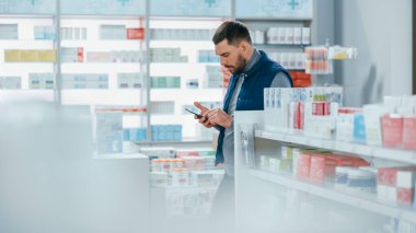 Eczane: Akıllı Telefon Kullanan Yakışıklı Beyaz Adam 'ın Portresi, En İyi İlaç, İlaç, Vitamin Almaya Çalışıyor. Raflar dolusu sağlık, sağlık ve spor malzemeleri.