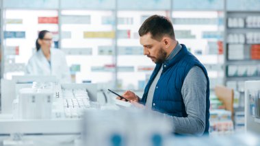 Eczane: Akıllı Telefon Kullanan Yakışıklı Beyaz Adam 'ın Portresi, En İyi İlaç, İlaç, Vitamin Almaya Çalışıyor. Raflar dolusu sağlık, sağlık ve spor malzemeleri.