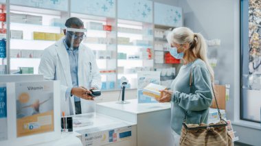 Eczane kasası: Yüz Koruyucu Giyen Profesyonel Siyah Eczacı, Yüz Maskesi Takan Kıdemli Kadın Müşteriye İlaç Satıyor, Temassız Ödeme Kullan. Coronavirus Güvenliği