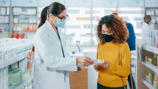 Covid-19 Yüz Maskesi Giyen Eczane: Profesyonel Eczacı Güzel Kadın Müşteriye Tıbbi Tavsiyeyle Yardım Ediyor, Konuşuyor. İlaç, hap, sağlık hizmeti ve güzellik ürünleriyle dolu bir eczane.