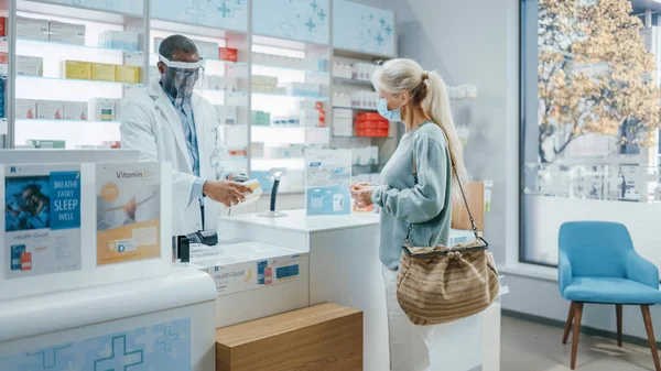 Pharmacy Drugstore Checkout Counter: Professional Black Pharmacist Wearing Face Shield Sells Medicine to Senior Female Customer, who is wearing Face Mask, Use Contactless Payment. Coronavirus Safety