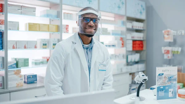 Pharmacy: Professional Confident Black Pharmacist Wearing Lab Coat and Face Shield, Looks at Camera Smiling Charmingly. Druggist in Drugstore Store with Shelves Medicine. Coronavirus Safety Measures