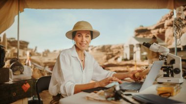 Arkeolojik Kazı Alanı: Kamerada Gülümseyen ve Dizüstü Bilgisayar Kullanan Ünlü Kadın Arkeolog, Fosil Antik Medeniyet Kültür Eserlerini Analiz Ediyor. Tarihçiler ekibi kazı alanında çalışıyor.