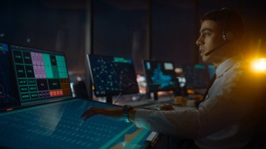 Havalimanı Kulesi 'nde Kulaklık Konuşması yapan Erkek Hava Trafik Kontrolörü. Ofis Odası, Takım için Navigasyon Ekranları, Uçak Gidiş ve Gelen Veri ile Masaüstü Bilgisayarı ile Doludur.