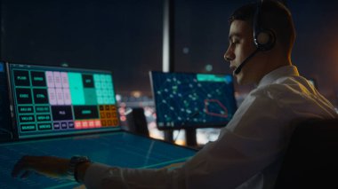 Havalimanı Kulesi 'nde Kulaklık Konuşması yapan Erkek Hava Trafik Kontrolörü. Ofis Odası, Takım için Navigasyon Ekranları, Uçak Gidiş ve Gelen Veri ile Masaüstü Bilgisayarı ile Doludur.