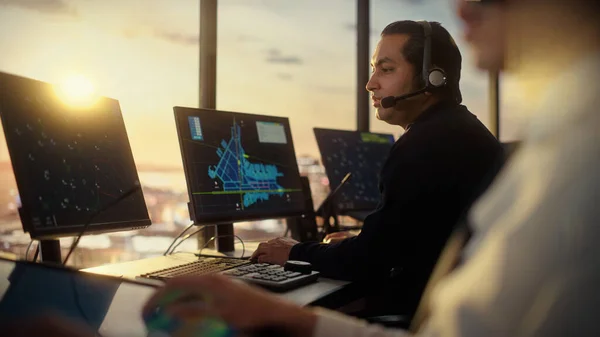Male Air Traffic Controller with Headset Talk on a Call in Airport Tower. Office Room is Full of Desktop Computer Displays with Navigation Screens, Airplane Departure and Arrival Data for the Team.