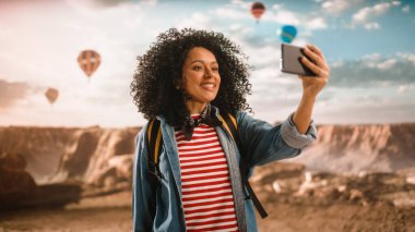 Afro Saç Tarzı ile Heyecanlı Genç Çok Irklı Gezgin Akıllı Telefon 'da Selfie Çekiyor ve Rocky Canyon Vadisi' nde Doğanın Güzelliğini Gösteriyor. Mountain National Park 'ta Sıcak Hava Balonu Festivali.