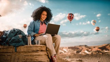 Rocky Canyon Vadisi 'nin tepesinde Afro Saç Tarzı Bilgisayarlı Heyecanlı Genç Çok Irklı Kadın Gezgin. Sıcak Hava Balonları Ulusal Dağ Parkı 'nda Uçuyor. Doğa yürüyüşü.