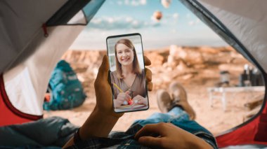 Bir Turistin POV 'u, Mutlu Gülümseyen Kız Arkadaşı ya da İş Arkadaşına Video Araması İçin Bir Çadırda Yatıyor ve Akıllı Telefon Kullanıyor. Gezgin Bir Dağın Tepesinde Dinleniyor Ve Uçan Sıcak Hava Balonları.