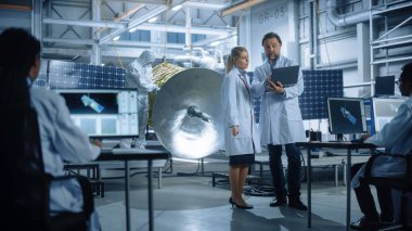 Mühendis ve teknisyen sohbeti. Uydu inşaatında çalışırken dizüstü bilgisayar kullanın. Uzay Ajansı İmalat Tesisi: Bilim adamları Uzay Keşif Görevi İçin Uzay Aracı İnşa Ediyor, Birleştiriyor