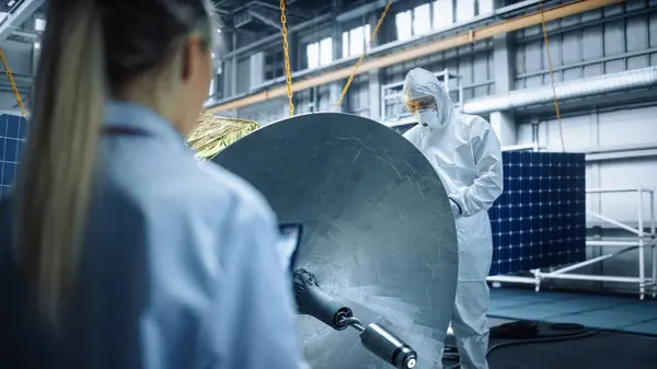 Female Engineer Uses Digital Tablet Computer while Working on Satellite Construction. Aerospace Agency: Scientists Assemble Spacecraft for Space Exploration Mission. Over the Shoulder Shot