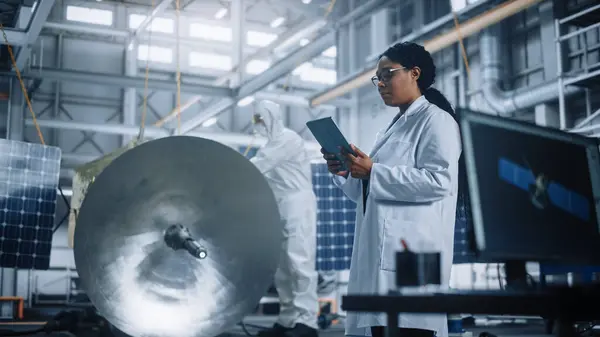 Female Engineer Uses Tablet Computer while Working on Satellite Construction. Aerospace Agency Manufacturing Facility: Scientists Build, Assemble Spacecraft for Space Exploration, Observation Mission