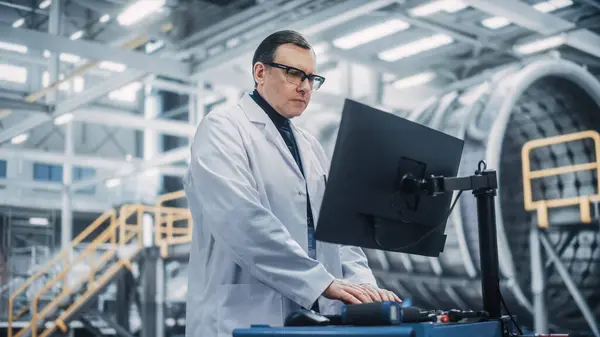 Professional Male Heavy Industry Engineer/Worker Working on a Computer. Confident Caucasian Industrial Specialist Wearing White Laboratory Coat and Safety Glasses in a Factory Facility.