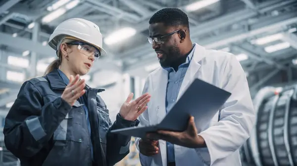 Team of Diverse Professional Heavy Industry Engineers Wearing Safety Uniform and Hard Hat Working on Laptop Computer. African American Technician and Female Worker Talking on a Meeting in a Factory.