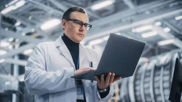 Professional Male Heavy Industry Engineer/Worker Working on a Laptop Computer. Confident Caucasian Industrial Specialist Wearing White Laboratory Robe in a Factory Facility.