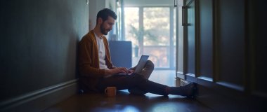 Evde Uzaktan Çalışmak İçin Bilgisayarı Kullanan Orta Yaşlı Adamın Portresi. Yerde oturan yakışıklı adam interneti kullanıyor, kahvenin sabahını içiyor. Üretim Çalışmalarının Olgun Atmosferi, Sosyal Medya Kullanımı