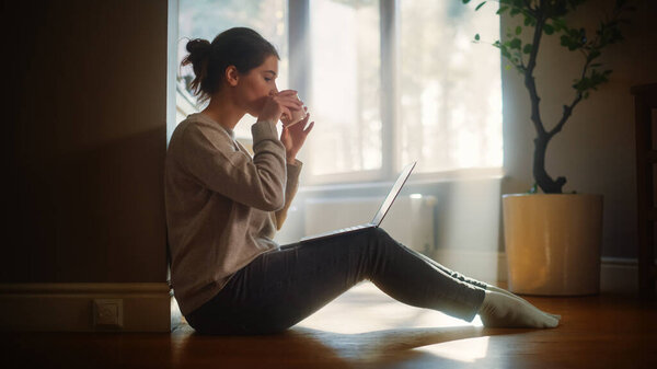Молодая женщина использует ноутбук дома для удаленной работы. Beautiful Girl Sitting on the Floor Использует Интернет, Напитки Утренняя чашка кофе. Солнечное утро благоприятно для продуктивной дистанционной работы