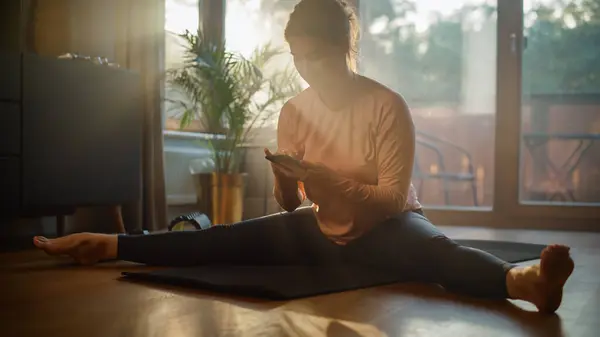 Beautiful Girl Using Smartphone to Help with Fitness Workout at Home. Smiling Woman Using Fitness Tracker, Yoga Exercises Application, Browsing Online Health Articles, Posting on Internet
