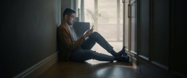 Evde Akıllı Telefon Kullanan Genç Yetişkin Adamın Portresi. Sırtı Duvara dayalı bir şekilde Koridor zemininde oturuyor. Web 'i Kontrol ve Sohbet İçin İnternet Kullanımı, Sosyal Medya Eğlencesi.