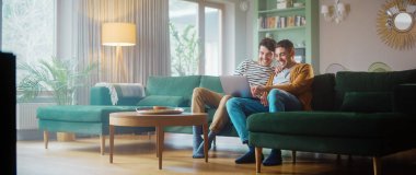 Konforlu Moda Apartmanında Kanepede otururken Dizüstü bilgisayar kullanan Nazik Gay Çiftin Portresi. Yetişkin Erkek Arkadaşları İnternet 'te Alışveriş, Canlı Servis' te Komik Videolar İzleme.