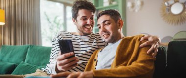 Cozy Stylish Apartmanında bir koltukta otururken akıllı telefon kullanan kibar eşcinsel çiftin portresi. Yetişkin Erkek Arkadaşları İnternet 'te Alışveriş, Canlı Servis' te Komik Videolar İzleme.