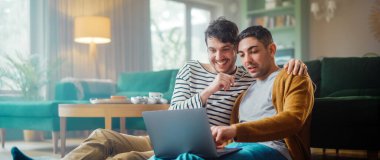 Yakışıklı Eşcinsel Çift Dizüstü bilgisayar kullanıyor, Oturma Odası zemininde otururken şık bir dairede. Yetişkin Erkek Arkadaşları İnternet 'te Alışveriş, Canlı Servis' te Komik Videolar İzleme.