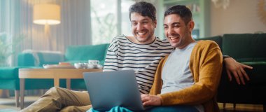 Yakışıklı Eşcinsel Çift Dizüstü bilgisayar kullanıyor, Oturma Odası zemininde otururken şık bir dairede. Yetişkin Erkek Arkadaşları İnternet 'te Alışveriş, Canlı Servis' te Komik Videolar İzleme.