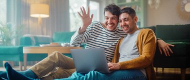 Yakışıklı Eşcinsel Çift Dizüstü bilgisayar kullanıyor, Oturma Odası zemininde otururken şık bir dairede. Yetişkin Erkek Arkadaşları Arkadaşları ya da Ailesi ile Çevrimiçi Video Görüşmesi Yapıyor.