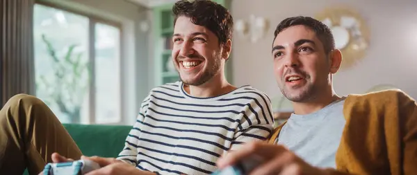 Happy Gay Pari Istuu Sohvalla Videopelien Pelaaminen Käyttäen Ohjaimia Kilpailukykyinen — kuvapankkivalokuva
