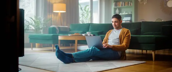 英俊的成年人使用笔记本电脑 而坐在舒适时尚公寓的客厅地板上 迷人的男人在网上购物 在流媒体上观看有趣的视频 — 图库照片