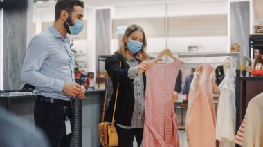 Giysi Mağazası: Kadın Müşteri Alışverişi, Perakende Satış Asistanı Yardım. Koruyucu yüz maskeleri takıyordu. Moda Dükkanındaki İnsanlar, Şık Kıyafetler Seçimi, Sürdürülebilir Tasarımlı Renkli Markalar