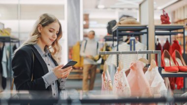 Güzel Gülümseyen Kadın Müşteri Alışverişi, Akıllı Telefon Kullanımı, İnternet Taraması, İnternette Karşılaştırma, Şık Kıyafetler Seçme. Moda Dükkanı, Renkli Markalar, Sürdürülebilir Tasarımlar