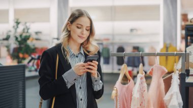 Güzel Gülümseyen Kadın Müşteri Alışverişi, Akıllı Telefon Kullanımı, İnternet Taraması, İnternette Karşılaştırma, Şık Kıyafetler Seçme. Moda Dükkanı, Renkli Markalar, Sürdürülebilir Tasarımlar