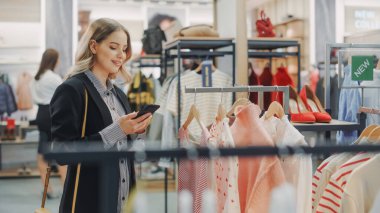 Güzel Gülümseyen Kadın Müşteri Alışverişi, Akıllı Telefon Kullanımı, İnternet Taraması, İnternette Karşılaştırma, Şık Kıyafetler Seçme. Moda Dükkanı, Renkli Markalar, Sürdürülebilir Tasarımlar