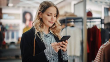 Güzel Gülümseyen Kadın Müşteri Alışverişi, Akıllı Telefon Kullanımı, İnternet Taraması, İnternette Karşılaştırma, Şık Kıyafetler Seçme. Moda Dükkanı, Renkli Markalar, Sürdürülebilir Tasarımlar